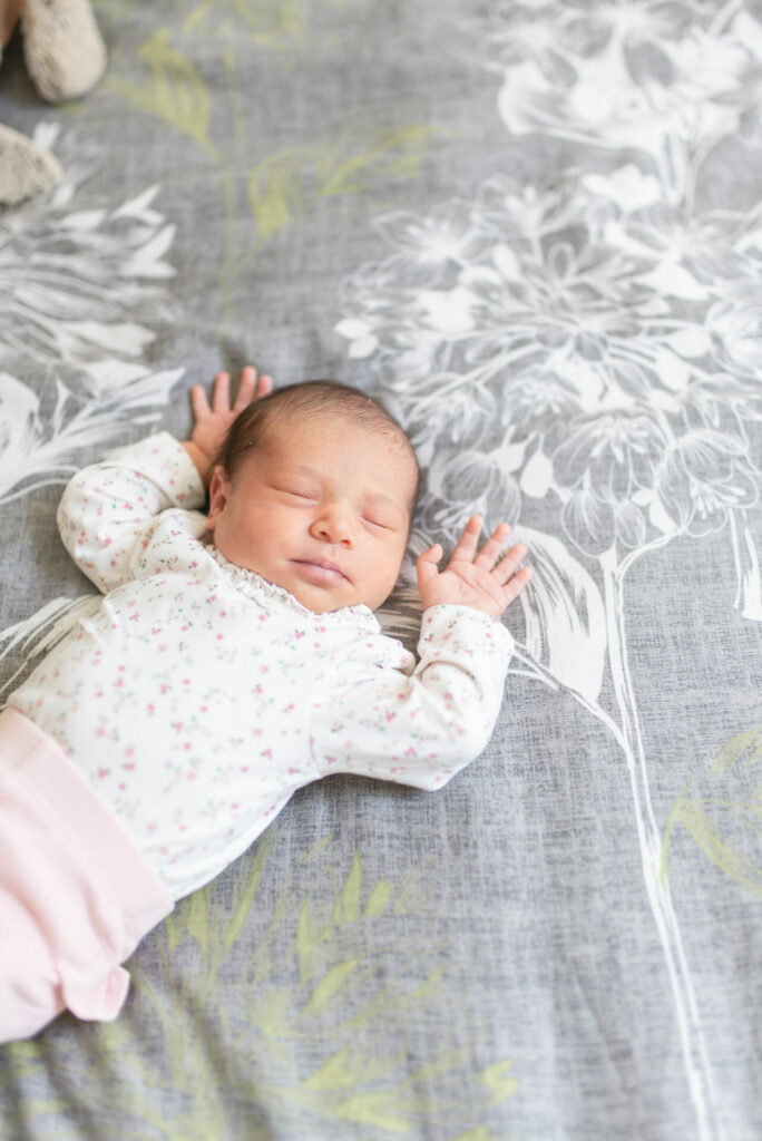 A baby lying on their back on a grey comforter, eyes closed and arms raised to their head.
